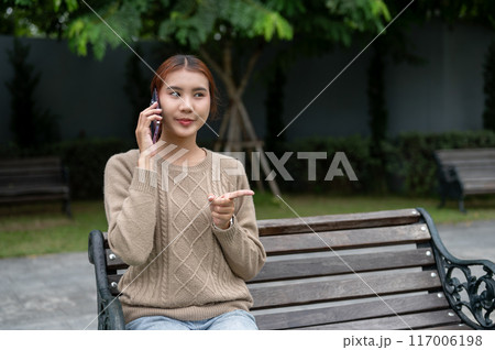 A beautiful Asian woman is talking on the phone with someone while resting on a bench in a park. 117006198