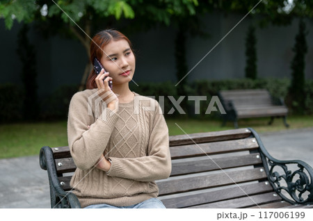 A beautiful Asian woman is talking on the phone with someone while resting on a bench in a park. 117006199