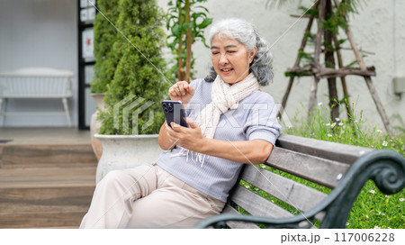 A positive, happy retired Asian woman is using her smartphone while relaxing in her home backyard. 117006228