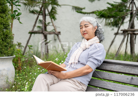 A calm mature Asian woman is sitting on a bench, her eyes closed and deep in thought while reading. 117006236