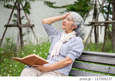 A mature Asian woman is feeling a headache and dizziness, while reading a book in her home backyard. A mature Asian woman is feeling a headache and dizziness, while reading a book in her home backyard. 117006237