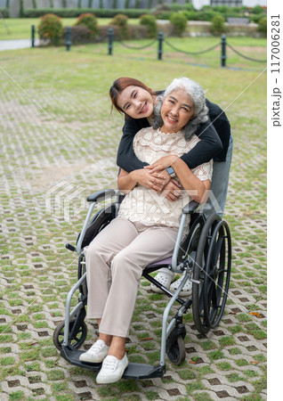 A lovely granddaughter is hugging her grandma, who is in a wheelchair, while walking her in a park. 117006281