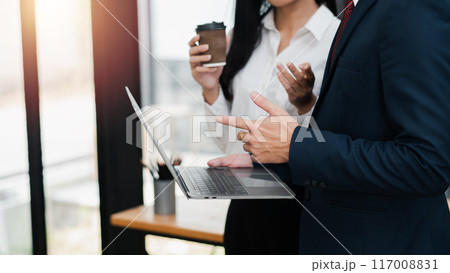 Two business professionals in formal attire discussing work while holding coffee cups in a modern office environment. Two business professionals in formal attire discussing work while holding coffee cups in a modern office environment. 117008831