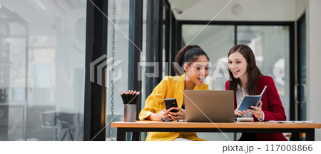 Two professional women working together in a modern office, using laptops and notebooks, smiling and discussing ideas. Two professional women working together in a modern office, using laptops and notebooks, smiling and discussing ideas. 117008866