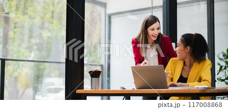 Two professional women working together on a laptop in a contemporary office setting with large windows and natural light. 117008868
