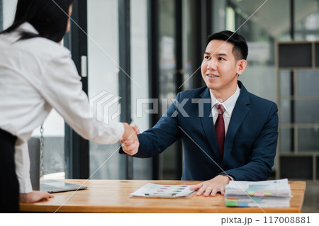 Two business professionals shaking hands across a table in a modern office environment, symbolizing agreement and partnership. 117008881