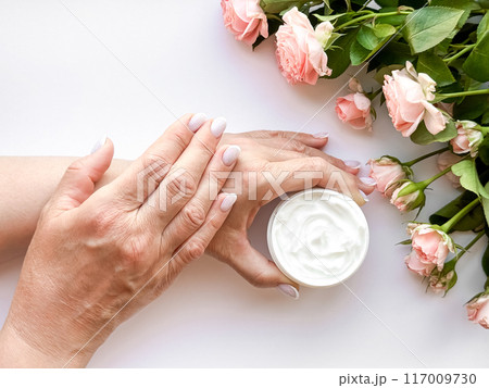 Hands middle age woman hold an open jar of white hand or body cream next to pink tea roses on white background, top view. Flat lay composition. Skincare and beauty product concept. 117009730