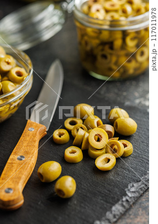 Sliced pickled green olives in brine on cutting board on black table. 117011978