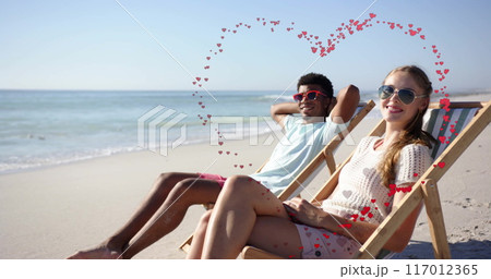 Image of hearts moving over diverse couple in love in deckchairs on beach in summer 117012365