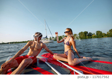 Young people, man and woman in swimwear and sunglasses enjoying sunny day on sup boards on river. Paddle boarding activity Young people, man and woman in swimwear and sunglasses enjoying sunny day on sup boards on river. Paddle boarding activity 117017562