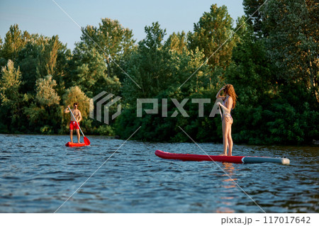 Active weekends. Young man and woman in swimwear enjoying summer day around nature, paddle boarding under clear blue sky. 117017642