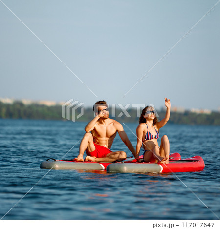 Young man and woman in swimwear and sunglasses sitting in the middle of the river on paddle boards and enjoying view 117017647