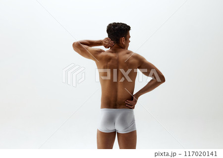 Rear view photo of fit young man in white boxer briefs stands with hand resting on hip and neck against white studio background. 117020141