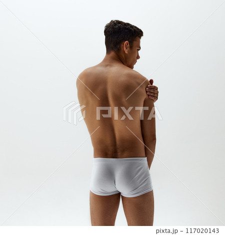 Rear view photo of fit young man in white boxer briefs stands with one hand resting on his shoulder against white studio background. Rear view photo of fit young man in white boxer briefs stands with one hand resting on his shoulder against white studio background. 117020143
