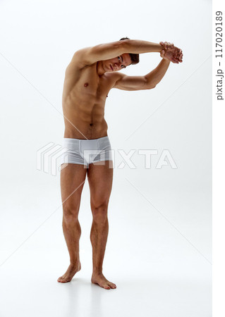 Young man in white boxer briefs stretching his back raising hands an doing bends against white studio background. 117020189