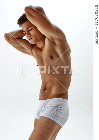 Muscular young man in white boxer briefs holds hands behind neck, highlighting his well-defined abs and chest against white studio background. Muscular young man in white boxer briefs holds hands behind neck, highlighting his well-defined abs and chest against white studio background. 117020218