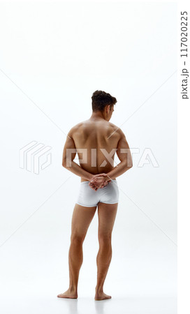 Rear view photo of fit man in boxer briefs stands holds hands behind his back, highlighting his toned physique against white studio background 117020225