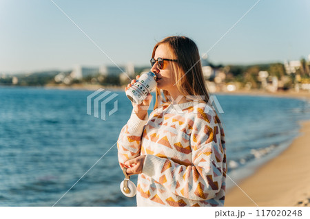 Young beautiful woman in cozy sweater and sunglasses enjoying hot coffee from thermo mug while relaxing on winter seaside sand beach. Cute attractive girl drinks tea taking sunbathe near ocean 117020248