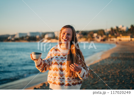 Young beautiful girl in cozy sweater holding coffee cup and smiling into the camera on seaside shore during mild sunset. Cute attractive woman enjoying her cup tea with autumn ocean waves background Young beautiful girl in cozy sweater holding coffee cup and smiling into the camera on seaside shore during mild sunset. Cute attractive woman enjoying her cup tea with autumn ocean waves background 117020249