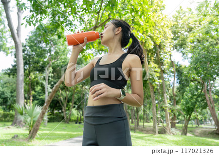 Fit Young Woman Drinking Water During Outdoor Workout in a Lush Green Park on a Sunny Day Fit Young Woman Drinking Water During Outdoor Workout in a Lush Green Park on a Sunny Day 117021326