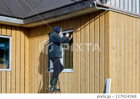 Diy installing metal window jamb and metal window stool in window opening from facade side, homeowner works standing on steel folding ladder. 117024568