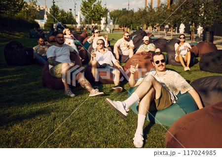 Group of happy young people, friends sitting on beanbag chairs, drinking beer, having fun and laughing. Outdoor picnic, festival Group of happy young people, friends sitting on beanbag chairs, drinking beer, having fun and laughing. Outdoor picnic, festival 117024985
