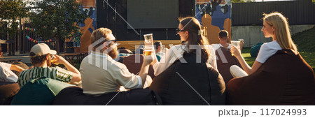 Group of people, men and women, friends gathering outdoor to watch movie on warm summer day, drinking beer and relaxing Group of people, men and women, friends gathering outdoor to watch movie on warm summer day, drinking beer and relaxing 117024993
