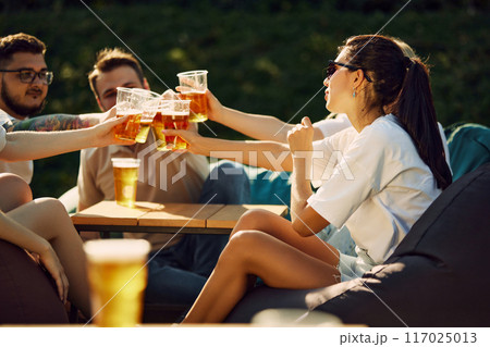 People meeting for a picnic, friends sitting together and drinking beer, clinking glasses, sharing joy and good conversations 117025013