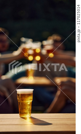 Focus on lager beer glass standing on wooden table with blurred people clinking glasses on background. Picnic Focus on lager beer glass standing on wooden table with blurred people clinking glasses on background. Picnic 117025014
