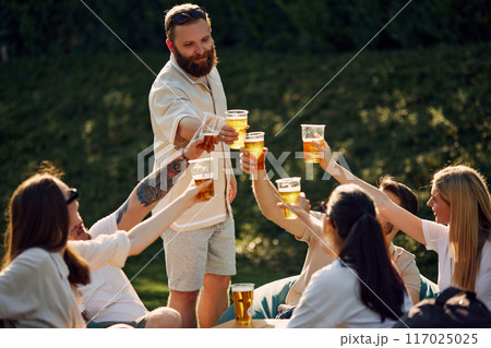 Group of happy smiling young people, friends gathering on outdoor picnic, drinking refreshing drink, having good company Group of happy smiling young people, friends gathering on outdoor picnic, drinking refreshing drink, having good company 117025025