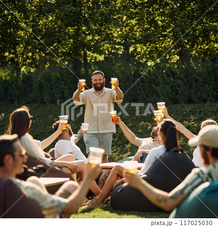 Group of happy smiling young people, friends gathering on outdoor picnic, drinking refreshing drink, having good company Group of happy smiling young people, friends gathering on outdoor picnic, drinking refreshing drink, having good company 117025030