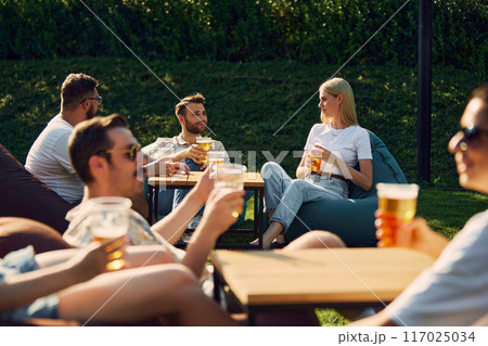Positive young men and women,friends gathering outdoors in public place, enjoying company, conversation and beer 117025034