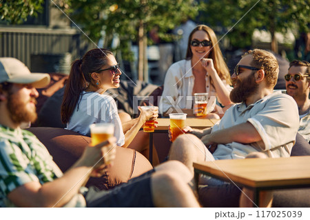 Lively Picnic. Young people, men and women, friends gathering on outdoor restaurant space, cheerfully talking and drinking beer. 117025039