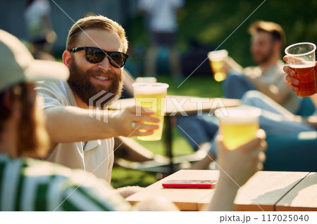 Sunny Celebration. Group of friends, gathering for a picnic, sharing laugh and engaging conversation while clinking beer glasses. 117025040