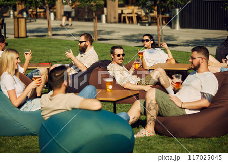 Cheerful young people, friends, men and women gathering on outdoor cafe space, for picnic, enjoying beer and conversations Cheerful young people, friends, men and women gathering on outdoor cafe space, for picnic, enjoying beer and conversations 117025045