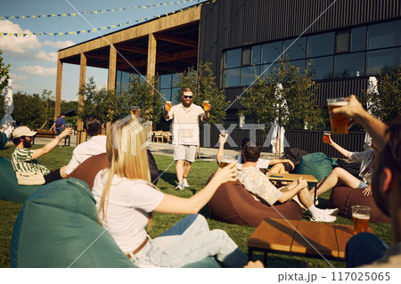 Happy young people, friends gathering in outdoor cafe space, having outdoor picnic with refreshing beer and cheerful talks 117025065