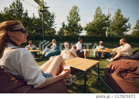 Chilled vibes. Friend, men and women lounging on beanbag chairs, drinking beer, cheerfully talking. Outdoor summer picnic in public place Chilled vibes. Friend, men and women lounging on beanbag chairs, drinking beer, cheerfully talking. Outdoor summer picnic in public place 117025071