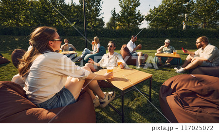 Group of young cheerful people, friends sitting on beanbag chair on grass, enjoying picnic, drinking beer, sharing joy and laugh Group of young cheerful people, friends sitting on beanbag chair on grass, enjoying picnic, drinking beer, sharing joy and laugh 117025072