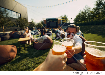 Cheers. Happy young people, friends gathering on outdoor picnic toasting foamy beer, clinking glasses. Refreshment of sunny hot days 117025073