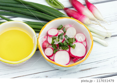 Salad of sliced radishes (French breakfast variety) and green onions in a little bowl, all ingredients with olive oil are on the white wooden rustic table. 117031479