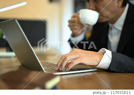Select focus on senior businessman typing on laptop keyboard while drinking coffee at desk Select focus on senior businessman typing on laptop keyboard while drinking coffee at desk 117031613