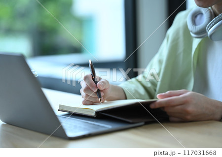 Close up shot of man sitting in front of laptop and handwriting important information on notepad Close up shot of man sitting in front of laptop and handwriting important information on notepad 117031668