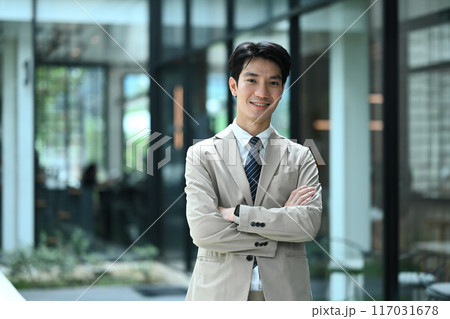 Portrait of smiling businessman standing outside modern office building with arms crossed Portrait of smiling businessman standing outside modern office building with arms crossed 117031678