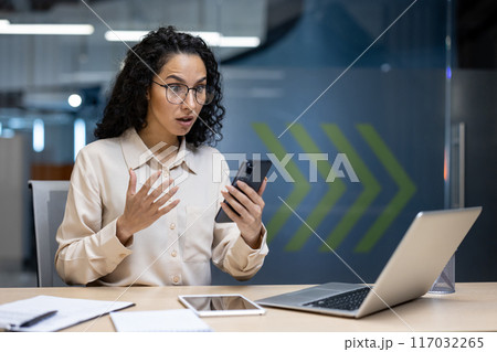 Shocked businesswoman looking at smartphone in office. Woman reacting to surprising or alarming news. Workspace with laptop, tablet, and notepad. Indoor professional setting. 117032265