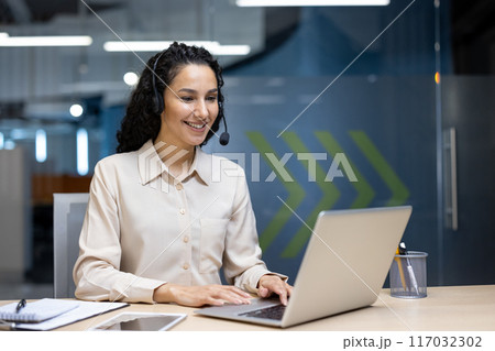 Smiling customer service representative wearing headset working on laptop in modern office environment. Woman engaged in online communication. Professional setting with documents, tablet Smiling customer service representative wearing headset working on laptop in modern office environment. Woman engaged in online communication. Professional setting with documents, tablet 117032302