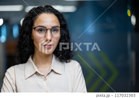 Portrait of confident businesswoman with glasses standing in modern office setting. Professional demeanor and serious expression convey leadership and professionalism. Office backdrop with blue tones Portrait of confident businesswoman with glasses standing in modern office setting. Professional demeanor and serious expression convey leadership and professionalism. Office backdrop with blue tones 117032303