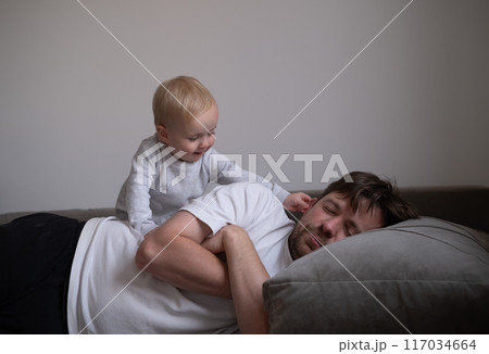 Cute little toddler girl walking over father, sleeping in bed in the morning. Cute little toddler girl walking over father, sleeping in bed in the morning. 117034664