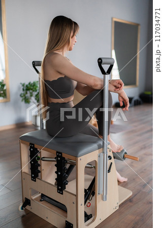 Portrait of Pilates fitness trainer sitting on Chair Balance Body machine in pilates studio. Young sporty attractive woman wearing sportswear in yoga studio. Fit, healthy and strong authentical body 117034771