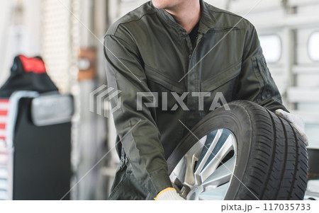 自動車整備工場でタイヤを運ぶ男性メカニック（肉体労働・重労働・重い・腰痛） 117035733