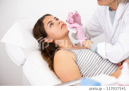 Professional mature female beautician with syringe injecting filler with hyaluronic acid in lips of young woman client during procedure in beauty salon 117037541